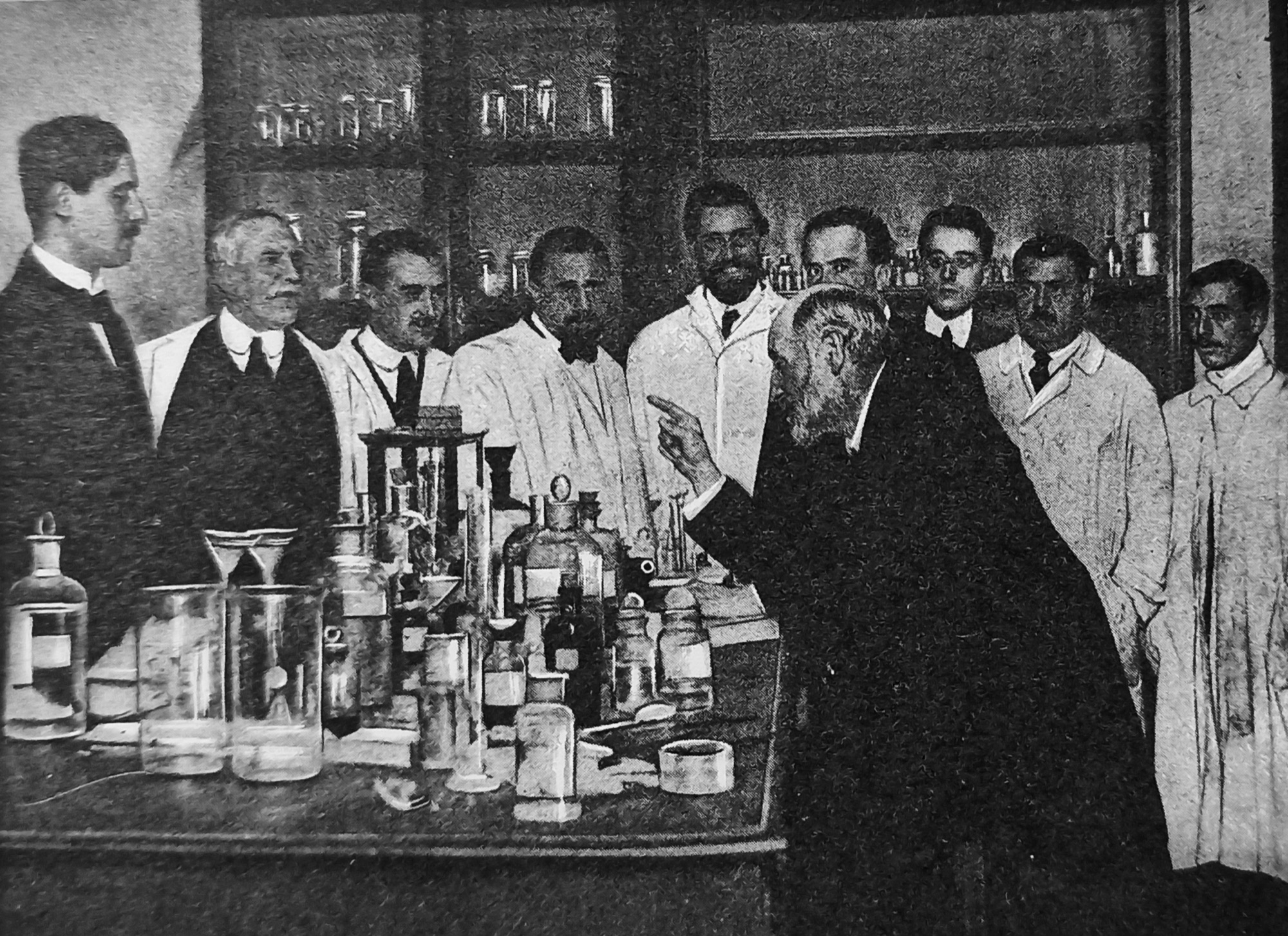 Santiago Ramón y Cajal pointing at a bench of glassware while students in lab coats watch.
